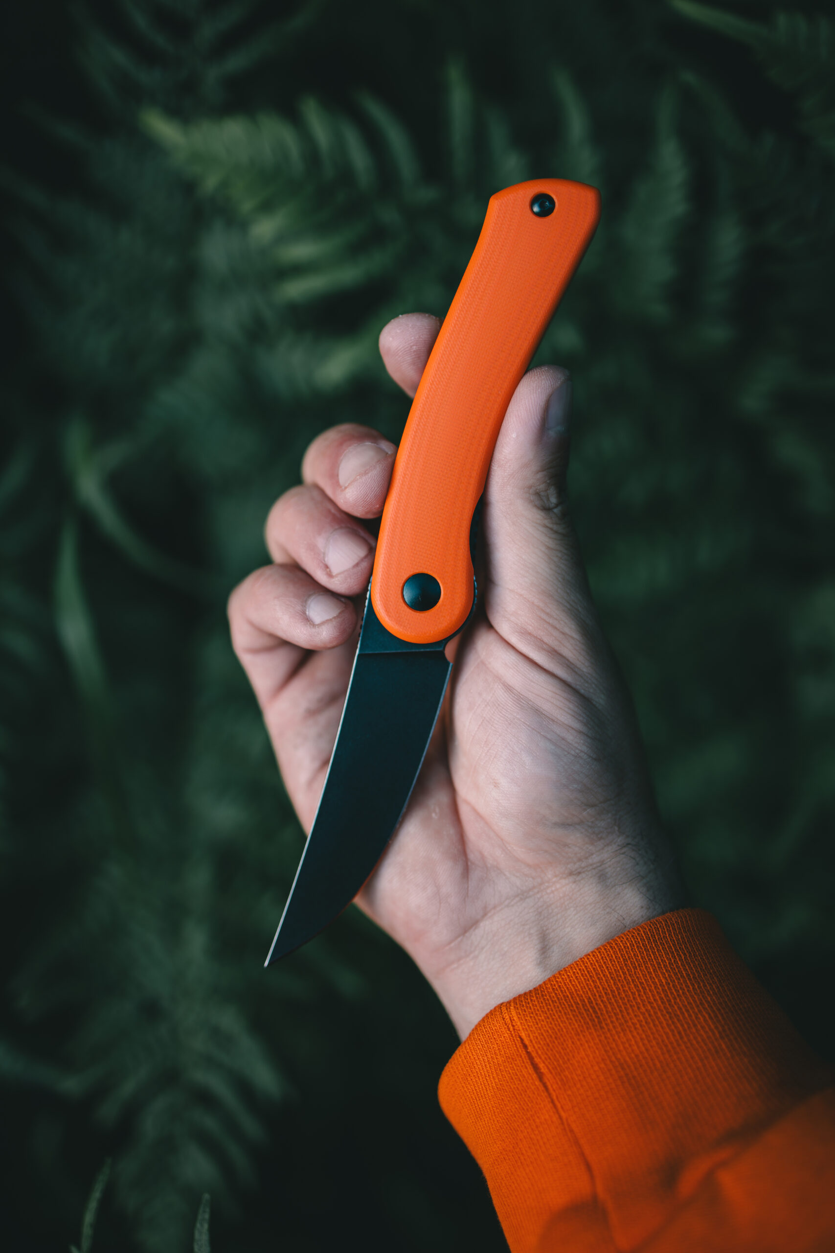 4 Hand holding a knife with an orange handle against a dark green leafy background